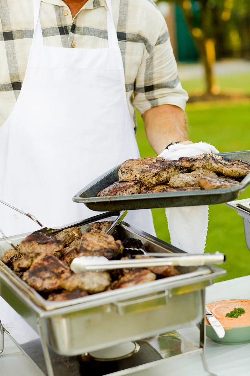 Man in white apron holding a tray of delicious grilled meats.