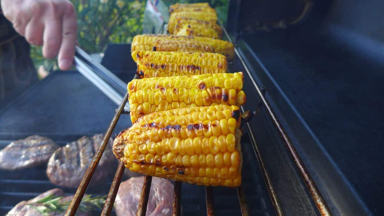 corn on the top shelf of a bbq grill with burgers below it and a hand with tongs