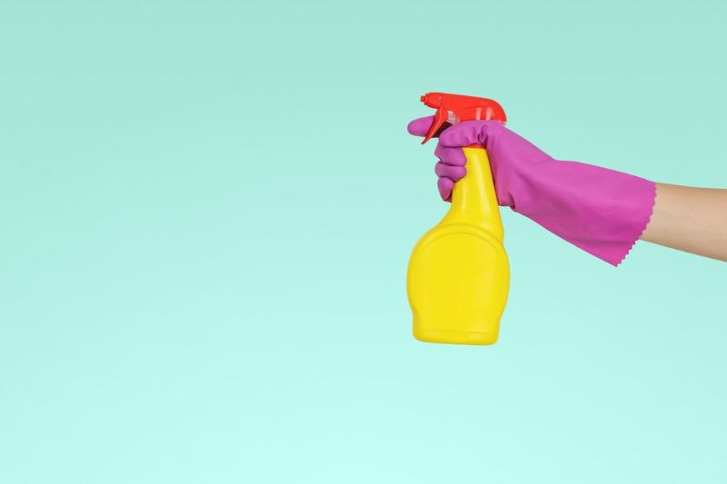 arm in a purple marigold glove holding a yellow and red spray bottle on a turquoise background
