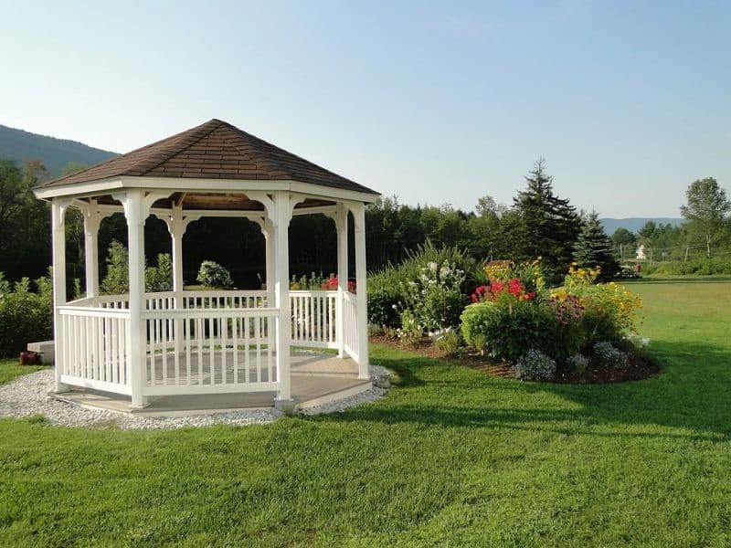 white gazebo bandstand on gravel patch by a lawn with bushes