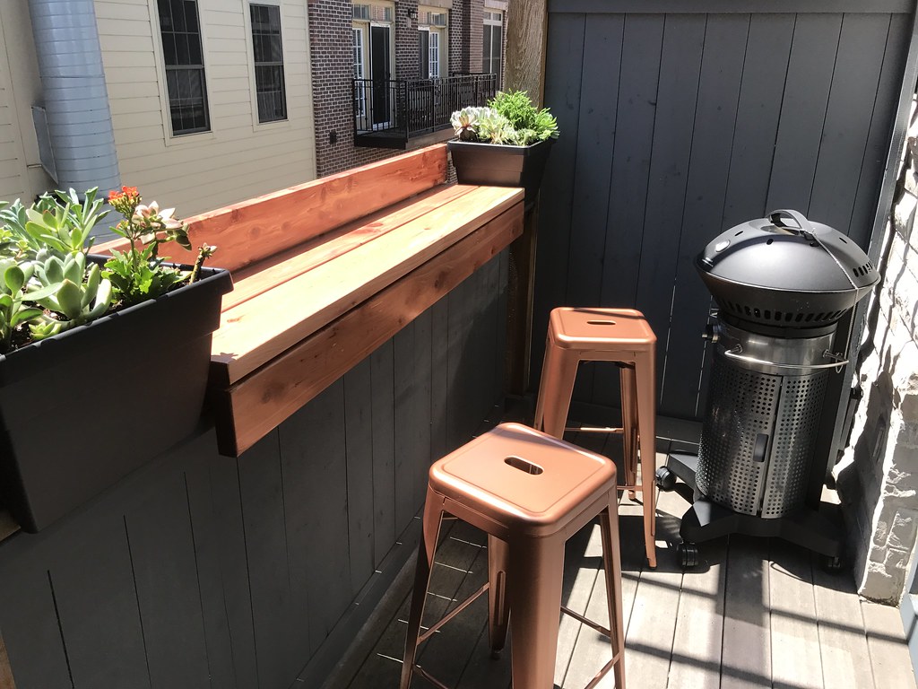 A small balcony setup with two metal stools, a wooden counter, potted succulents, and a modern outdoor heater.