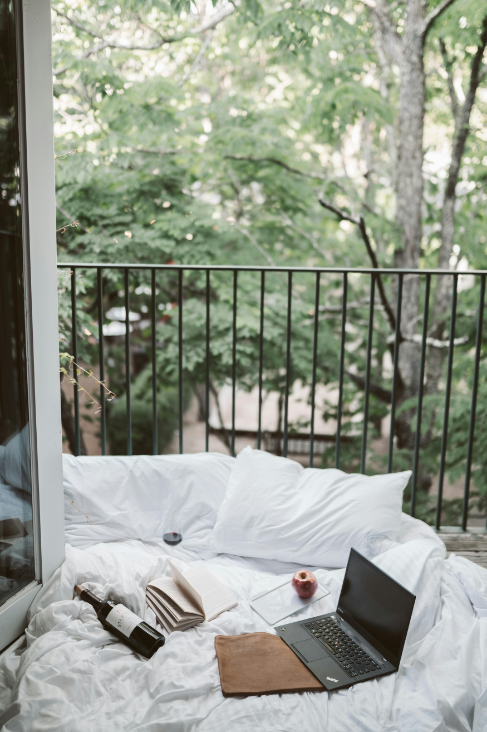 A cosy outdoor bed with pillows, a laptop, a book, and a bottle of wine on a balcony.