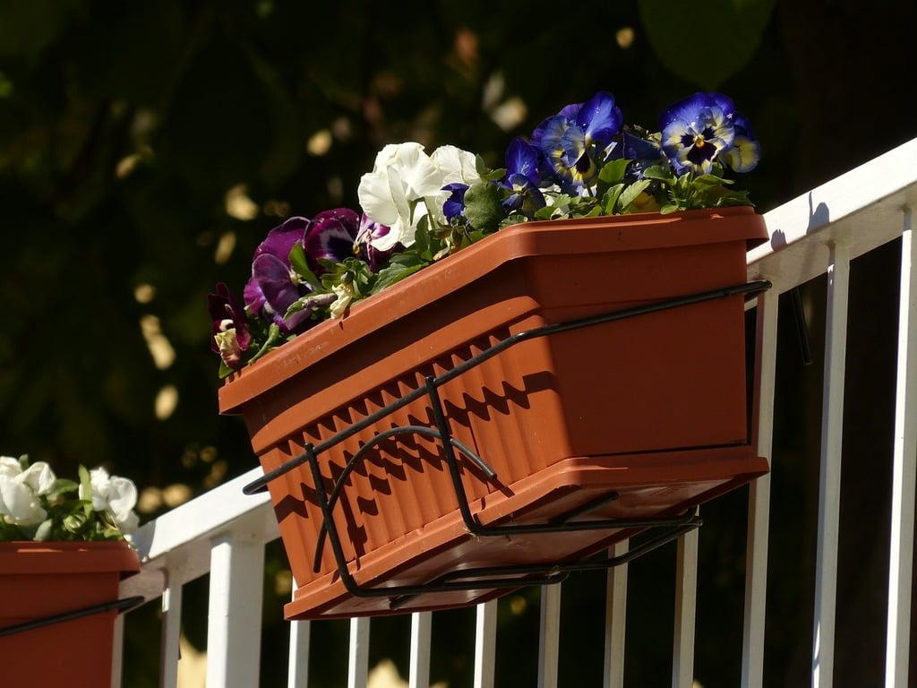 A rectangular plastic planter with purple and white flowers mounted on a white balcony railing.