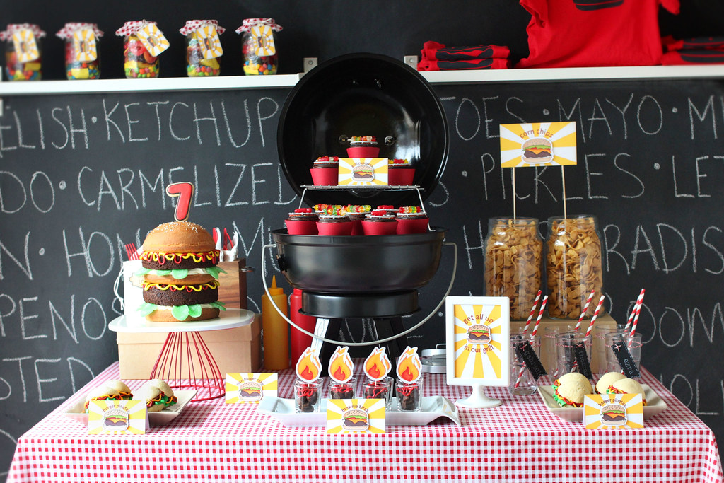 A table decorated with red Gingham tablecloth, with a hamburger cake on top, a faux grill filled with cupcakes, and more sweets
