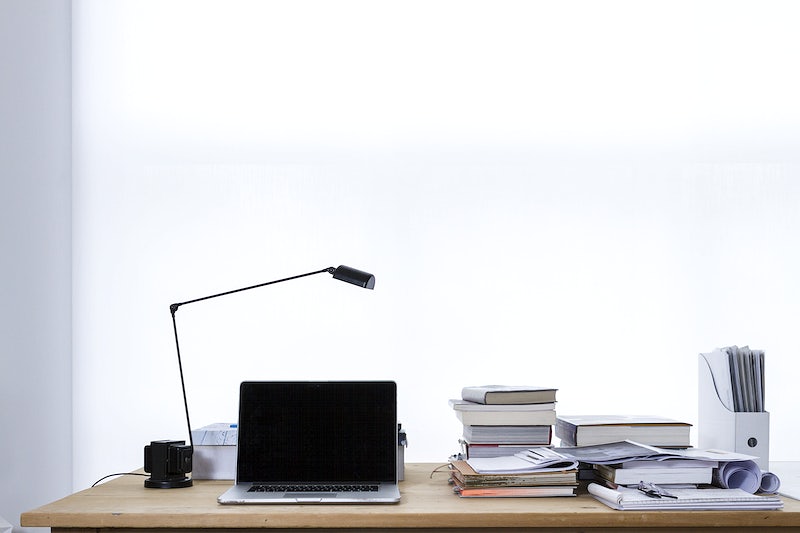 Laptop and lamp on desk with stacks of books and files