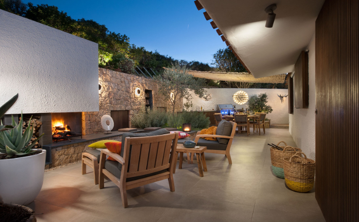 Multi-zone seating patio featuring wooden lounge chairs by a fireplace, a dining area with BBQ grill, potted plants, and stylish lighting.