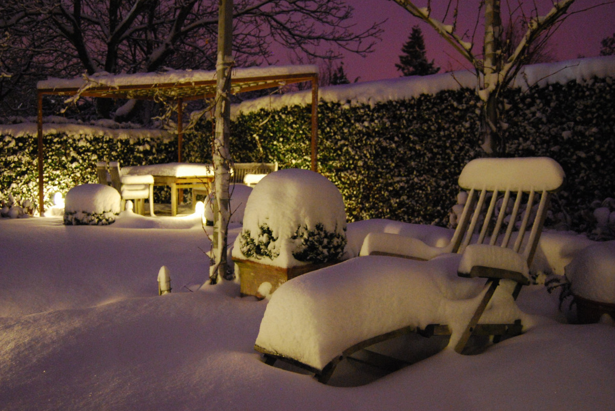 A snow-covered outdoor garden scene with chairs, a table, and plants blanketed in thick snow under evening light.