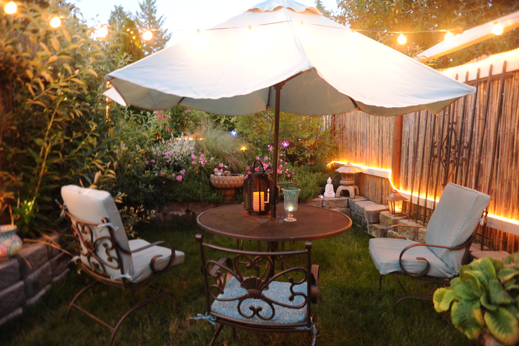 A cosy garden with a parasol, metal table, cushioned chairs, string lights, plants, flowers, and a small Buddha statue.