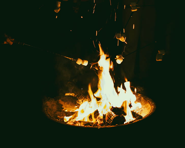 Several marshmallows on sticks being roasted over a fire in a round fire pit, with flames and sparks visible.