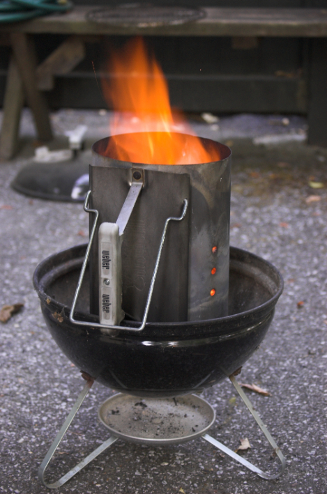 A lit charcoal chimney starter placed on a small barbecue grill, with flames coming from the top.