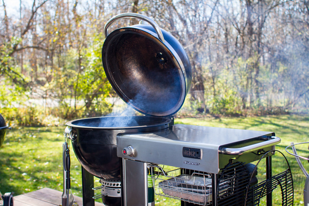 A charcoal smoker grill with its lid open, emitting smoke, placed outdoors on a sunny day.