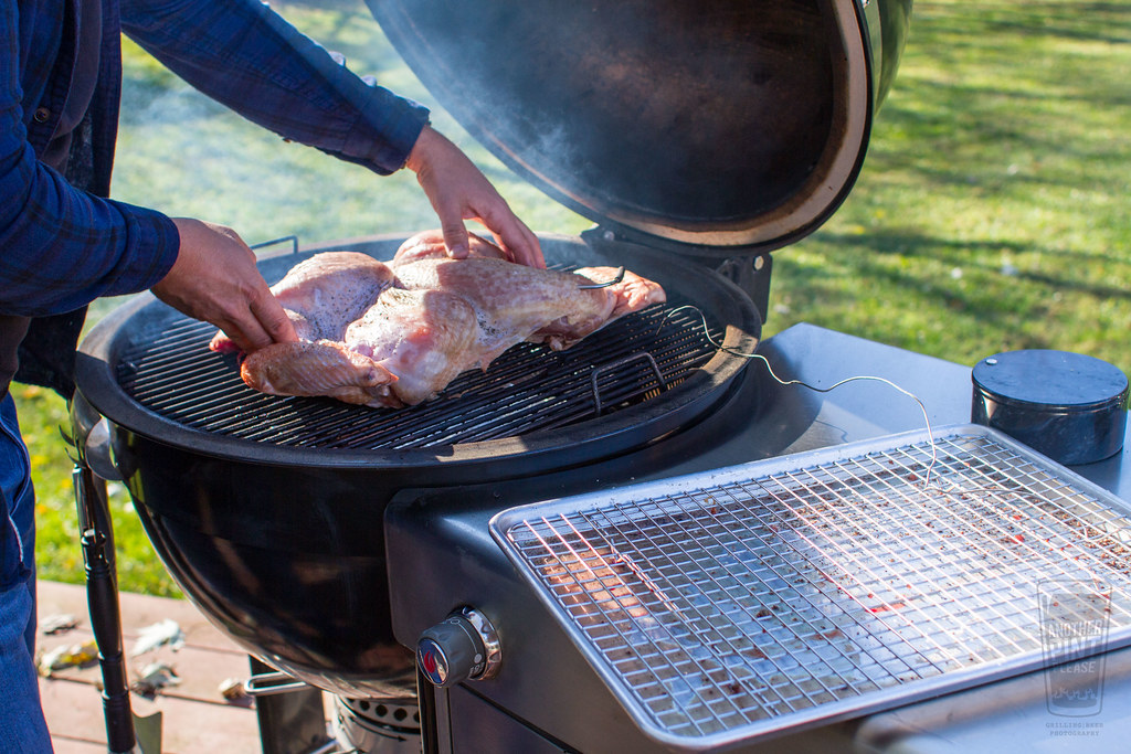 A person placing a raw whole chicken onto a smoking grill outdoors, with smoke rising from the grill.