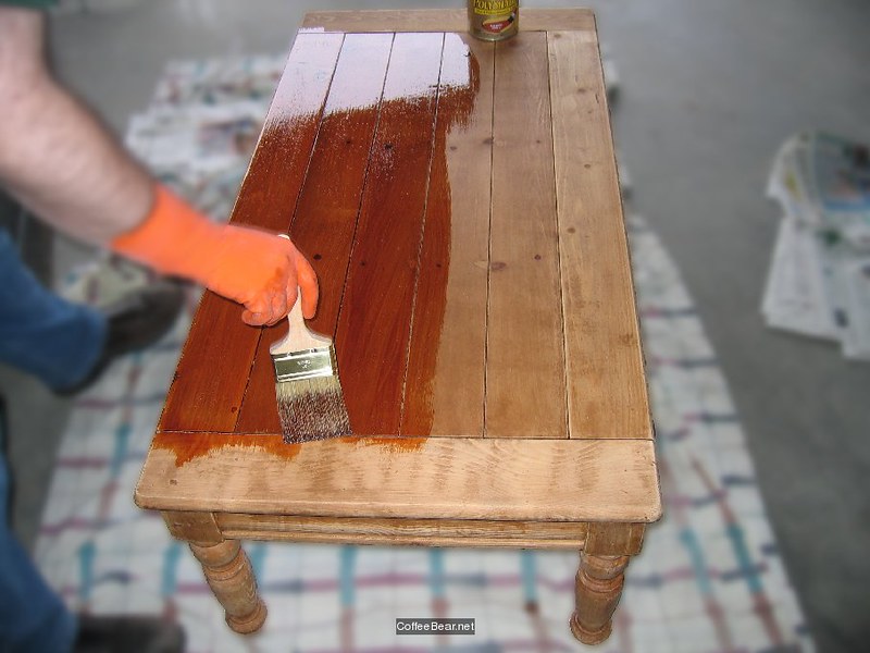 A person wearing orange gloves is applying wood sealant to a wooden table, with half stained and half unstained.