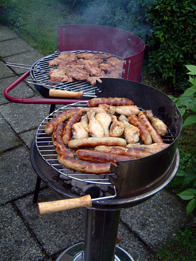 Two round grills with covers/barriers, cooking assorted meats like sausages and chicken outdoors on a patio with greenery nearby.