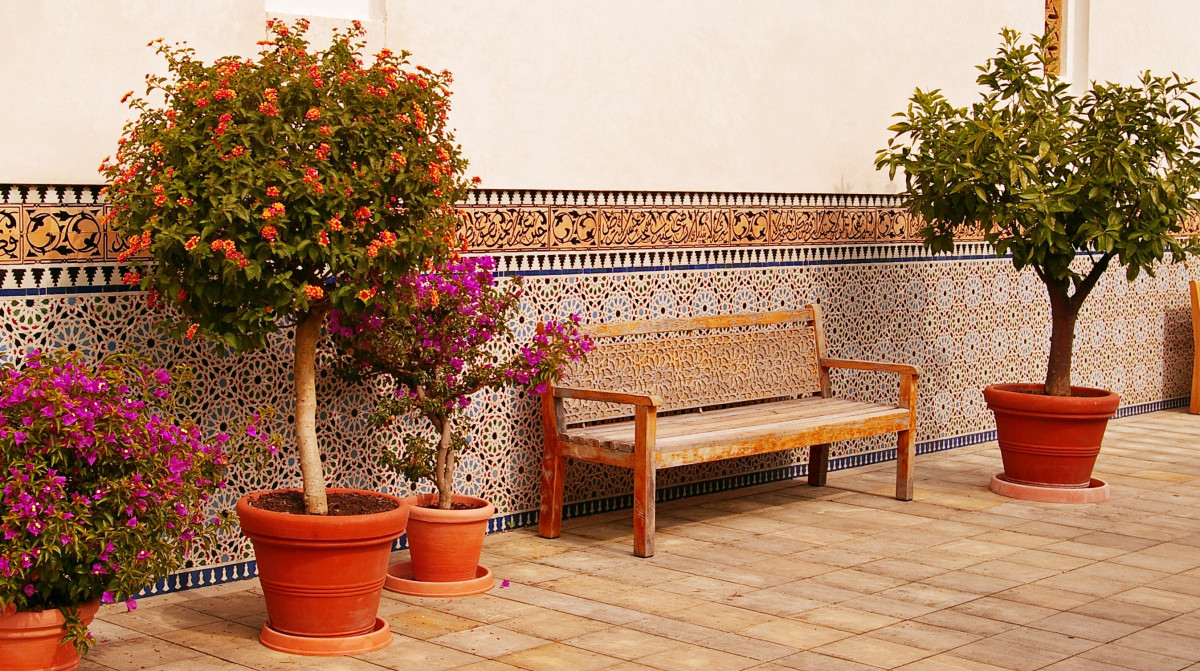 A Mediterranean-style courtyard featuring a terracotta colour scheme with vibrant potted plants, a wooden bench, and intricate patterned tiles.