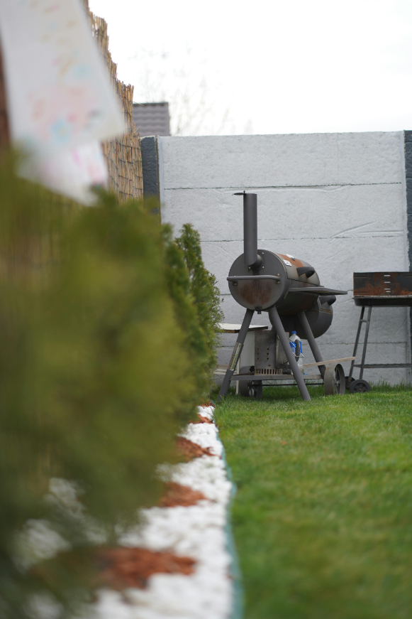  A backyard setup with a barbecue grill placed near a fence, surrounded by neatly trimmed bushes, white stones, and green grass.