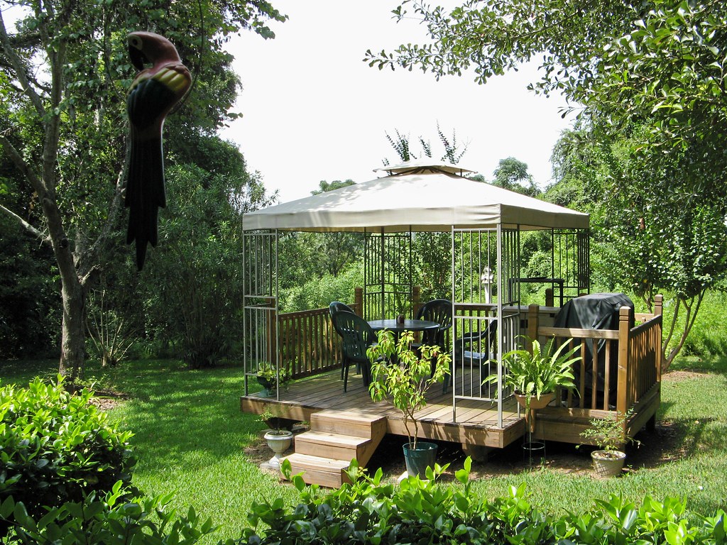 A garden gazebo with a small deck, canopy, table, chairs, and a barbecue grill, surrounded by lush greenery and plants.