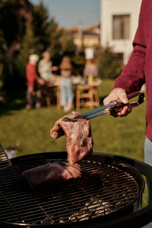 A person holding tongs with a large piece of raw meat over a grill, with a group gathering in the background.