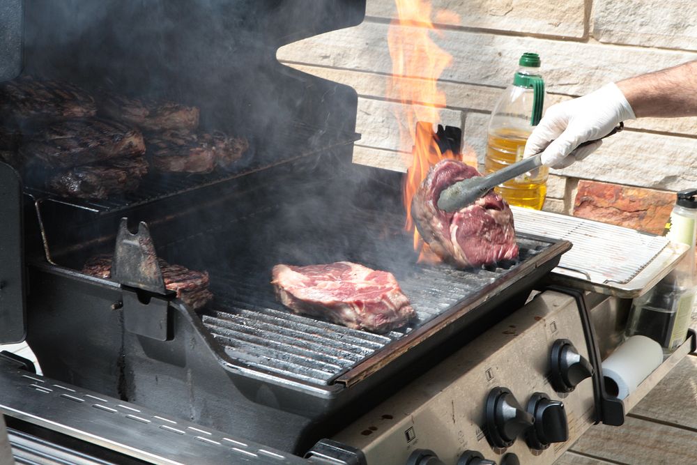 A person grilling steaks on a gas grill, with one steak being turned using tongs as flames rise from the grill.