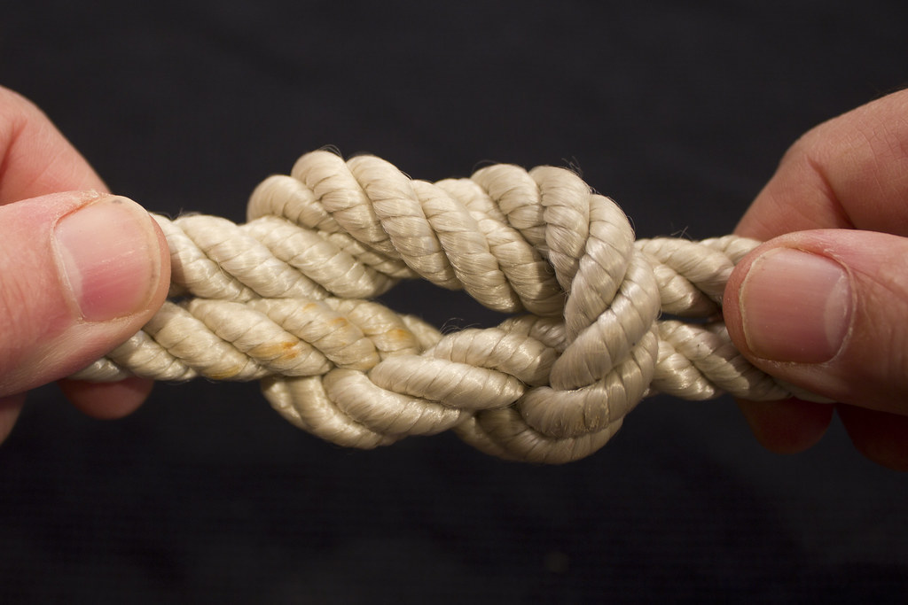 Close-up of hands holding a tied square knot in a thick rope against a dark background.