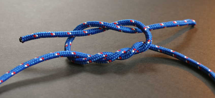 A close-up of a blue rope with red and white accents tied into a surgeon’s knot, lying on a dark surface