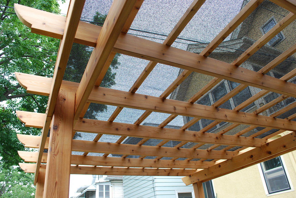 A wooden pergola with a lattice roof and a black mesh covering, providing shade to a backyard patio area.