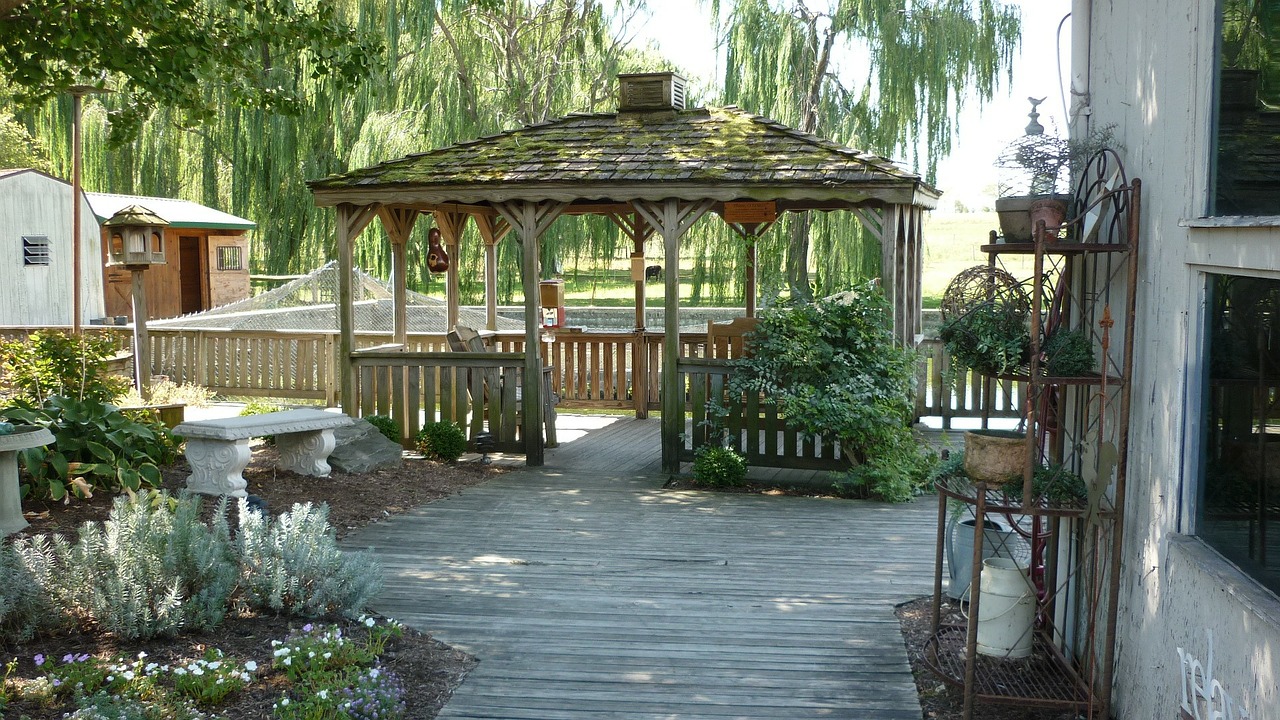 A rustic wooden pergoda with a shingled roof, surrounded by greenery and a garden, in a backyard setting.