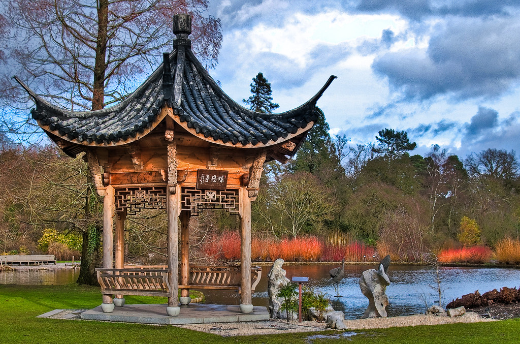 A traditional Chinese pagoda with an ornate, curved roof situated beside a serene pond in a landscaped garden.