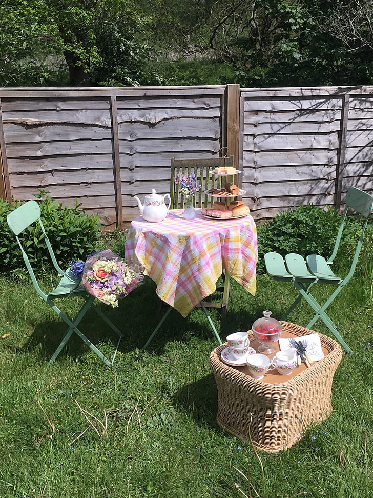 A cosy outdoor tea setup in a sunlit garden with a small table, chairs, teapot, tiered stand, and a wicker basket