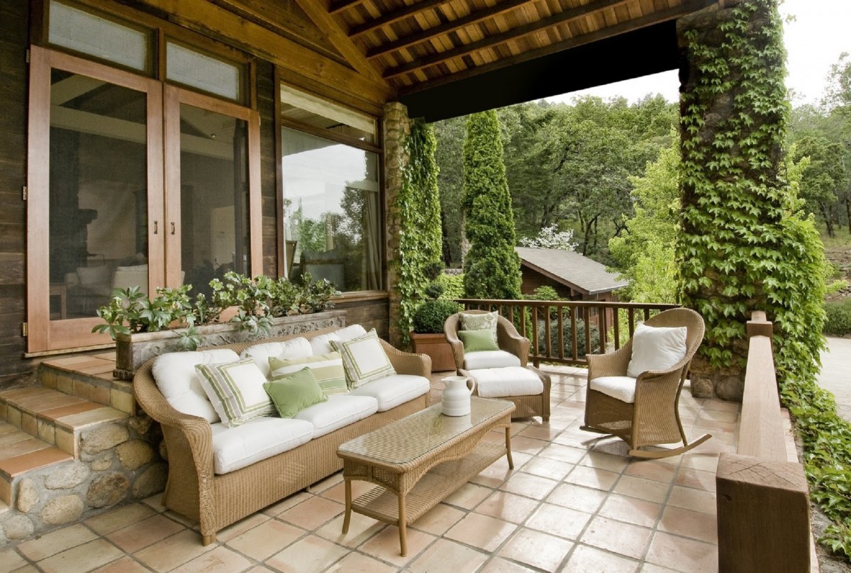 A shaded patio setup under a pergola with wicker furniture, green cushions, and potted plants, surrounded by lush greenery and ivy-covered columns.