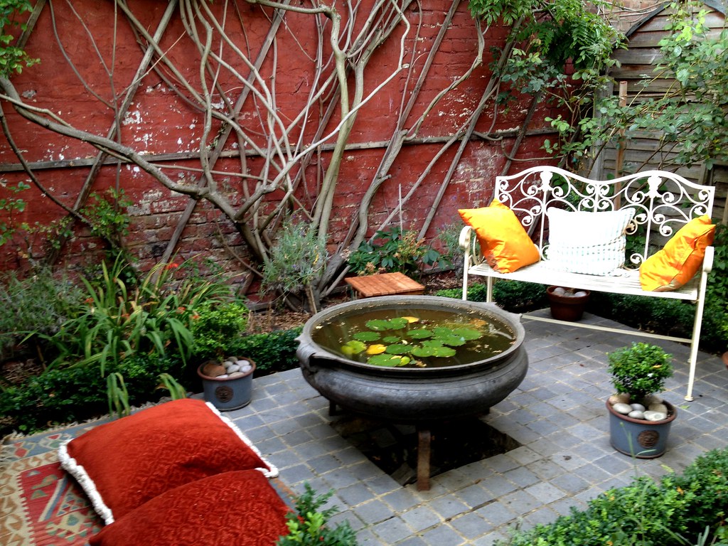 A garden nook tucked in the corner with a white metal bench, orange cushions, and a water feature surrounded by greenery.