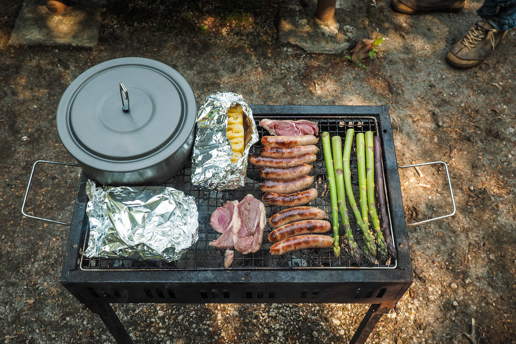 A camping grill with sausages, asparagus, and foil-wrapped food next to a pot, being cooked outdoors.