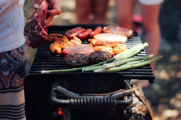 A person grilling various meats and asparagus on a barbecue grill outdoors, with others in the background.