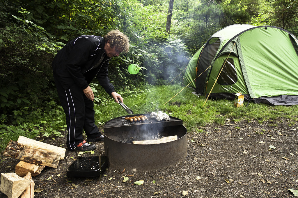 A man grilling sausages and foil-wrapped food at a campsite with a green tent in the background.