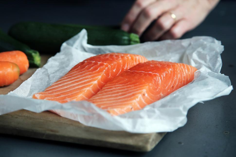 Two fresh salmon fillets on parchment paper, with carrots and zucchini in the background, prepared for cooking.