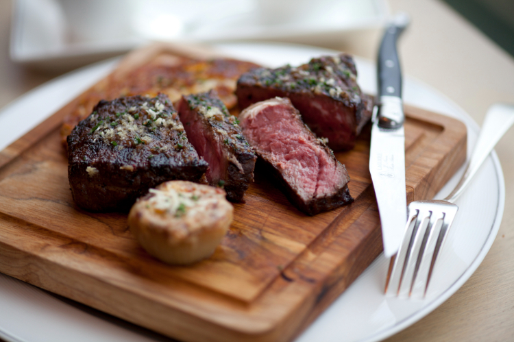 A sliced medium-rare steak on a wooden board, accompanied by a knife, fork, and a small side dish.