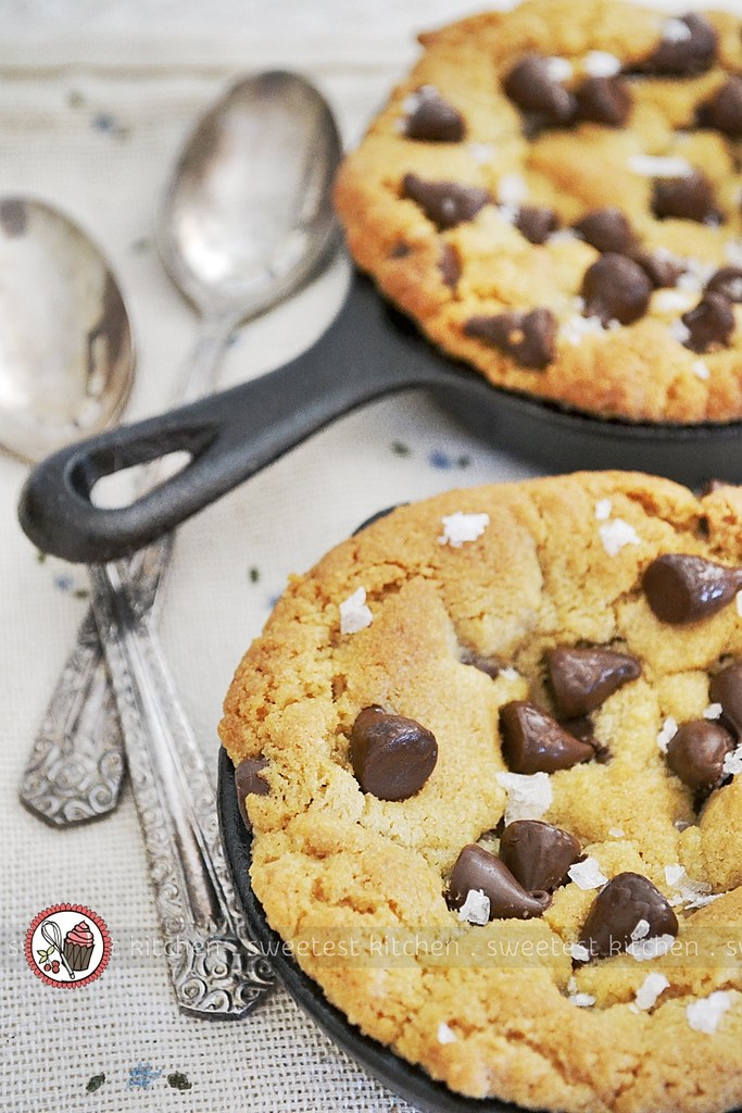 Two skillet-baked chocolate chip cookies topped with sea salt flakes, accompanied by vintage spoons placed on a tablecloth.