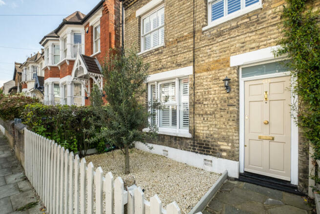 A terraced house with a small, fenced front garden featuring a gravel surface, an olive tree in the centre, and white-framed windows and door.