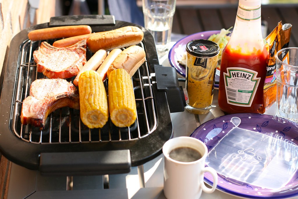 A portable gas grill set up on a balcony, with corn, buns, and meat on the grill.